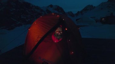 Kırmızı ceketli bir adam kamp çadırında oturuyor. Parlayan Noel çelengi ışıklarıyla pencereden bakıyor. Gece vakti kış dağları.