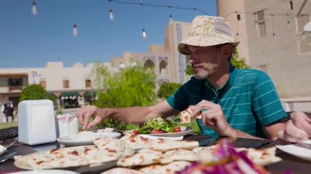 帽子の男の観光客は 中央アジアのウズベキスタンのブハラの古い町広場のレストランの国立プレートでウズベキスタンの料理とサムサまたはサンボエを食べています — ストック動画