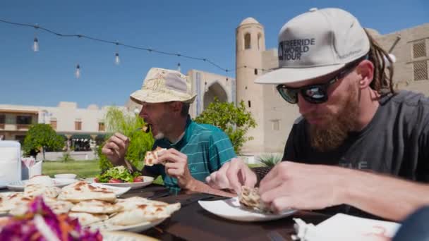 帽子をかぶった2人の男性観光客がウズベキスタンの料理を食べている サムサまたはサンボエは 中央アジアのウズベキスタンのブハラにある古い町の広場のレストランのナショナルプレートで — ストック動画