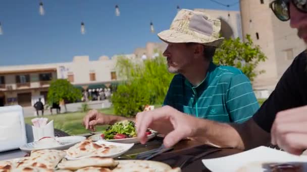 帽子をかぶった2人の男性観光客がウズベキスタンの料理を食べている サムサまたはサンボエは 中央アジアのウズベキスタンのブハラにある古い町の広場のレストランのナショナルプレートで — ストック動画