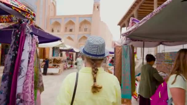 身穿黄色衬衫 头戴黄帽的年轻女子在尖塔和马德拉斯附近拥挤的街道市场上行走 乌兹别克斯坦Khiva的Itchan Kala古城 — 图库视频影像