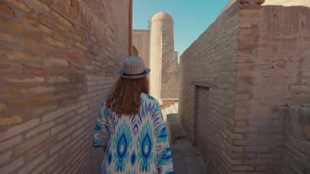 身着民族服装 头戴蓝色饰物和帽子的年轻女子 面对着马德拉斯的背景 走在古城狭窄的街道上 乌兹别克斯坦Khiva的Itchan Kala古城 — 图库视频影像
