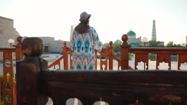 ブルーの装飾とモスクの近くの帽子とウズベキスタンのイッチャカラの古代都市キヴァの広場でミナレットの民族服の若い女性 — ストック動画