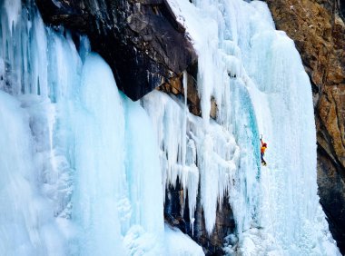 Turuncu ceketli sporcunun Barskoon 'daki büyük donmuş şelaleye tırmanışının insansız hava aracı görüntüsü Kırgızistan' daki dağ vadisini dolduruyor.