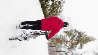 Kırmızı ceketli bir adamın karlı parkta bisikletiyle yürüdüğü dikey bir video. Kar kenti Almatı, Kazakistan 'da kış bisikleti