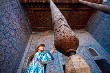 Woman tourist in ethnic dress touch wooden carved column at Inner Yard of blue mosaic palace hall in Tash Hauli of ancient city Khiva in Uzbekistan. clipart