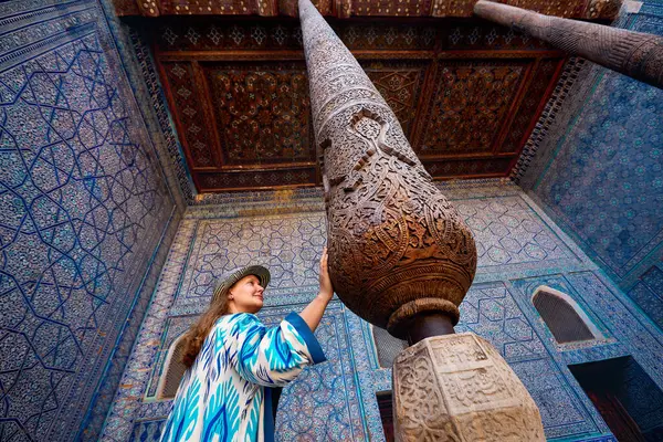 stock image Woman tourist in ethnic dress touch wooden carved column at Inner Yard of blue mosaic palace hall in Tash Hauli of ancient city Khiva in Uzbekistan.