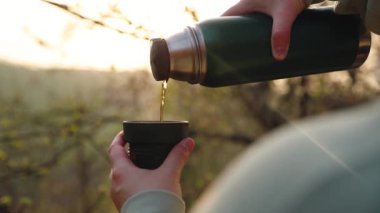 Kadın gezgin, gün batımında bahçede yeşil termostan sıcak çay dolduruyor.