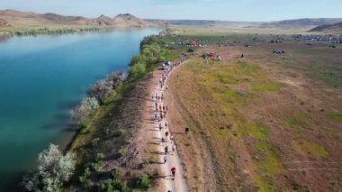 Güneşin doğuşunda nehir ve tepelerin yanındaki tozlu köy yolunda yarışan kalabalığın insansız hava aracı görüntüsü. Güney Kazakistan bozkırında ultra maraton koşusu