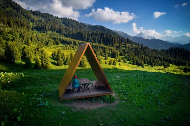 Mavi ceketli turist yürüyüşçüsü kadın, Almaty, Kazakistan 'daki vahşi dağ ve ladin ormanlarının güzel manzaralı hava aracının üçgen çatılı yazlığında dinleniyor.