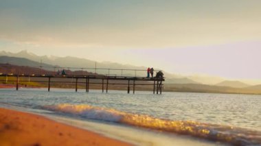 Issyk kul Gölü 'nde gün batımında iskelede balıkçılar ve arka planda dağlarla. Kırgızistan 'da dalgalar ve kumlu plaj