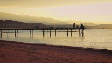 Issyk kul Gölü 'nde gün batımında iskelede balıkçılar ve arka planda dağlarla. Kırgızistan 'da dalgalar ve kumlu plaj