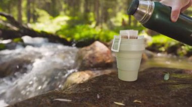 Yeşil bardakta kahve poşeti. Yaz ormanlarındaki dağ nehrinin yakınındaki kayalıklardaki termostan sıcak suyla demlenen içeceğin işlemleri..