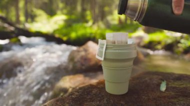 Yeşil bardakta kahve poşeti. Yaz ormanlarındaki dağ nehrinin yakınındaki kayalıklardaki termostan sıcak suyla demlenen içeceğin işlemleri..