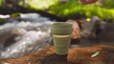 Kadın yeşil kupaya Drip Coffee Bag koymuş. Yaz ormanlarındaki dağ nehrinin yakınındaki kayalıklarda demlenen içecek işlemleri..