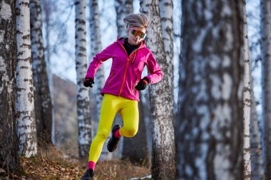 Pembe ceketli atletik koşucu kadın ve sarı eşofman altı ormanda patikada koşuyor.