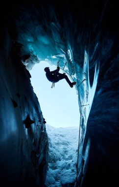 Siyah siluetli adam buz baltası ve kramponlarla buz dağlarındaki donmuş buz mağarasına tırmanıyor. Spor dağcılığı ve dağcılık konsepti.
