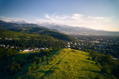 Hava aracı manzaralı yeşil tepe ve arka planda kar dağları manzaralı evler Kazakistan, Almaty şehrinde.