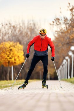 Sonbahar parkında kask ve kırmızı ceketli patenciler üzerinde antrenman yapan erkek sporcu. Kayak dağı disiplini için İskandinav kayak konsepti
