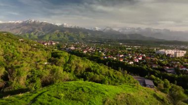 Hava aracı manzaralı evler yeşil tepe vadisi manzaralı kar dağları günbatımında gökyüzü arka planında Kazakistan, Almaty şehrinde