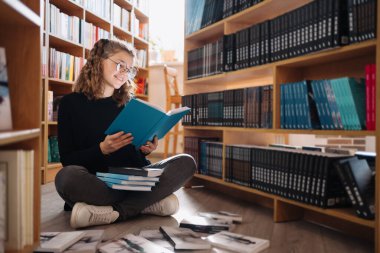 Bir yığın kitabın arasında genç bir kız. Gözlüklü genç bir kız arka planda raflar olan bir kitap okuyor. O, kitap yığınları ile çevrilidir. Kitap günü.