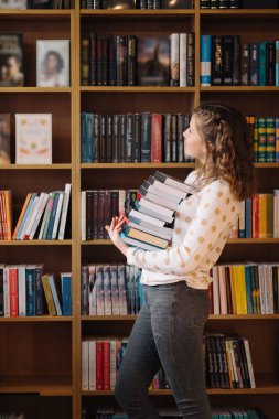 Teen girl among a pile of books. A young girl holding books with shelves in the background. She is surrounded by stacks of books. Book day.