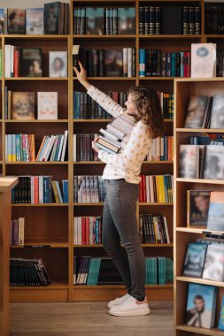 Teen girl among a pile of books. A young girl holding books with shelves in the background. She is surrounded by stacks of books. Book day.