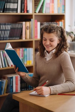 Attractive happy young girl student studying at the college library, sitting at the desk