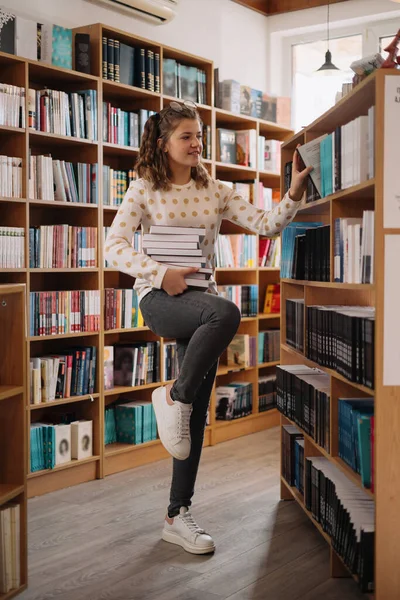 本の山の中で10代の女の子 背景に棚のある本を持っている少女 彼女は本の山に囲まれている 予約日 — ストック写真