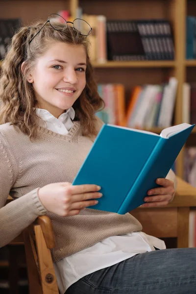 本の山の中で10代の女の子 若い女の子が背景に棚のある本を読んでいる 彼女は本の山に囲まれている 予約日 — ストック写真
