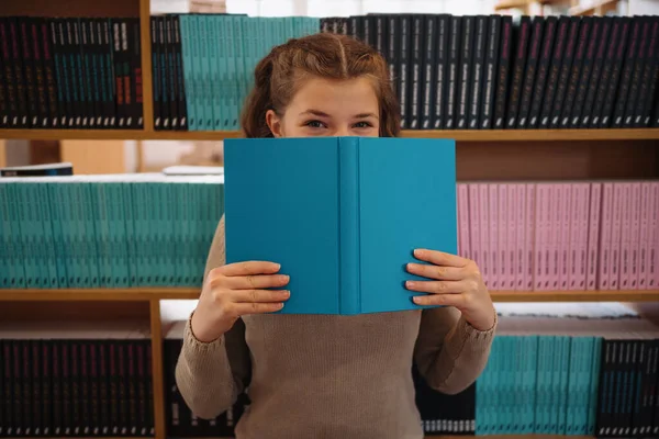 開いている青い本の後ろに隠れているかなり若い女の子の肖像画と見下ろします本棚の背景 — ストック写真