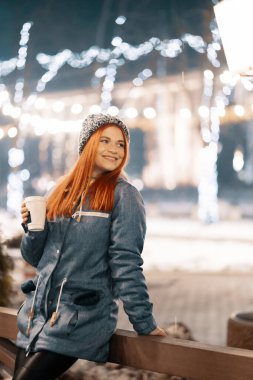 Girl drinking hot coffee while walking in the evening city market decorated with holiday lights in the evening. Feeling happy in big city. Spending winter vacations