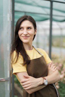 Smiling florist businesswoman portrait wearing yellow t-shirt and brown apron holding arms crossed at the green house