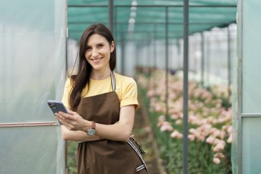 Smiling florist businesswoman using smartphone, wearing yellow t-shirt and brown apron holding phone in hands at the green house