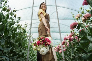 Woman florist walking among flowers in a green house carrying a basket with a fresh bouquet