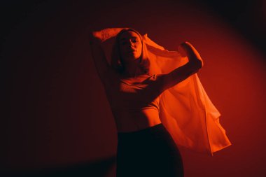 Lady in crop top raising trendy jacket and looking at camera against black backdrop in dim neon light