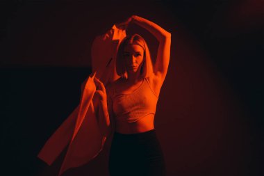 Lady in crop top raising trendy jacket and looking at camera against black backdrop in dim neon light