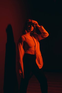 Serene female model in trendy jacket closing eyes while standing against black backdrop in dark studio