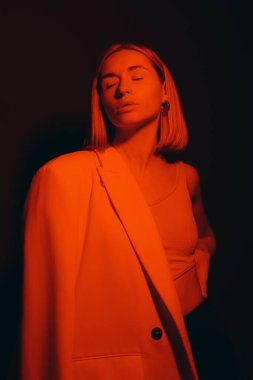 Serene female model in trendy jacket closing eyes while standing against black backdrop in dark studio