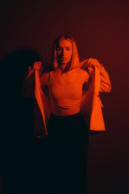Serene female model in trendy jacket closing eyes while standing against black backdrop in dark studio