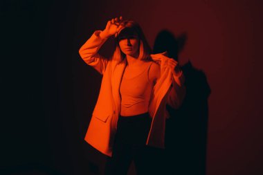 Serene female model in trendy jacket closing eyes while standing against black backdrop in dark studio