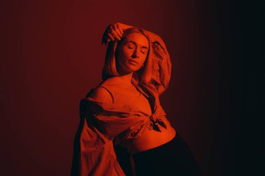 Relaxed blond lady in stylish outfit closing eyes and touching head while standing against red background in neon light