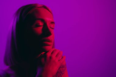 Dreamy female model closing eyes while praying for success against violet background in neon illumination