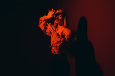 Relaxed blond lady in stylish outfit closing eyes and touching head while standing against red background in neon light