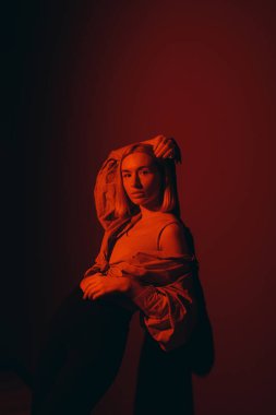 Relaxed blond lady in stylish outfit closing eyes and touching head while standing against red background in neon light