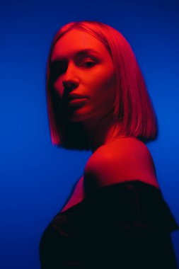 Young female with fair hair looking at camera against blue backdrop in studio with neon illumination