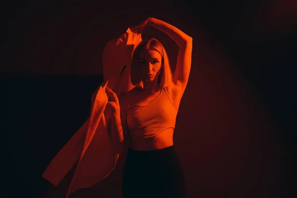 Lady in crop top raising trendy jacket and looking at camera against black backdrop in dim neon light