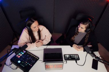 Female host and guest sitting at table with equipment and tablet with On Air inscription while broadcasting radio program in recording studio