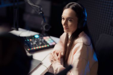 Young female with headphones clasping hands and looking away while sitting at table during podcast in recording studio