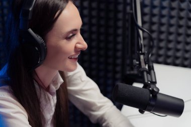 Happy brunette with headphones smiling and looking away while talking into microphone during radio broadcast in contemporary studio